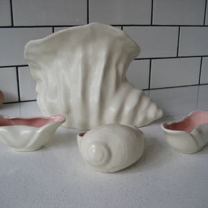 Peut inclure: Un ensemble de quatre bols en céramique blanche en forme de coquillage avec des intérieurs roses. Le plus grand bol est un vase avec une large ouverture et une base étroite. Les trois autres bols sont plus petits et ont une forme plus arrondie.