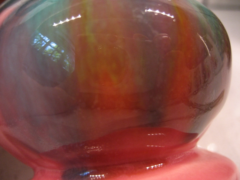 May include: A round, iridescent glass vase with a pink base. The vase has a swirling pattern of green, blue, and red.