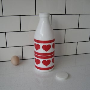 May include: White glass milk bottle with a red and white heart pattern. The bottle has a white lid.