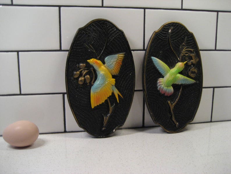 K&ouml;nnte beinhalten: Zwei schwarze Wandplaketten mit einem 3D-Design von V&ouml;geln. Eine Plakette zeigt einen gelben Vogel mit schwarzen und blauen Akzenten, w&auml;hrend die andere Plakette einen gr&uuml;n-blauen Vogel mit wei&szlig;en Akzenten zeigt.