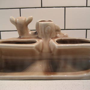 Może przedstawiać: Ceramiczna doniczka z trzema oddzielnymi przegr&oacute;dkami, z ozdobną figurką jelenia na g&oacute;rze. Doniczka jest jasnobrązowa z szkliwioną powierzchnią.