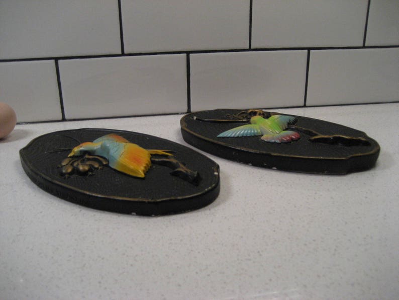 K&ouml;nnte beinhalten: Zwei schwarze und goldene Wandplaketten mit einem 3D-Kolibri-Design. Ein Kolibri ist gr&uuml;n, gelb und blau. Der andere Kolibri ist blau, gelb und orange.