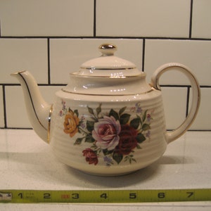 May include: A white teapot with a floral design featuring pink, red, and yellow roses. The teapot has a gold trim around the lid and spout. The teapot is 18 cm wide.