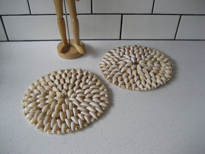 May include: Two round, white and beige, shell-shaped coasters. The coasters are made of many small shells that are arranged in a circular pattern.