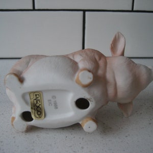 May include: A ceramic pig figurine in a lying down position, with a light pink and white color scheme. The figurine has a matte finish and features small, rounded legs and ears. The underside of the pig has a sticker that reads "Enesco" and "1988 EIC".