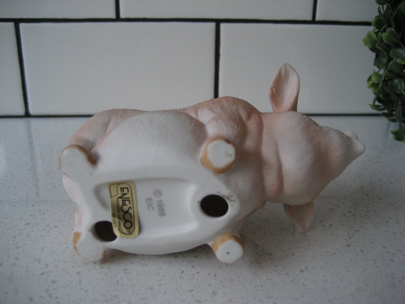 May include: A ceramic pig figurine in a lying down position, with a light pink and white color scheme. The figurine has a matte finish and features small, rounded legs and ears. The underside of the pig has a sticker that reads "Enesco" and "1988 EIC".