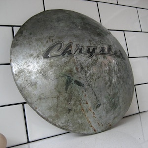 Puede incluir: Un tapacubos plateado y desgastado con la palabra "Chrysler" en relieve en letras negras.