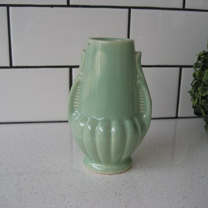 Puede incluir: Un pequeño jarrón de cerámica verde claro con dos asas. El jarrón tiene un diseño acanalado y un fondo redondeado.