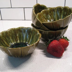 May include: Set of four green ceramic bowls with a scalloped edge. The bowls are stacked on top of each other. There are two red strawberries in the foreground.