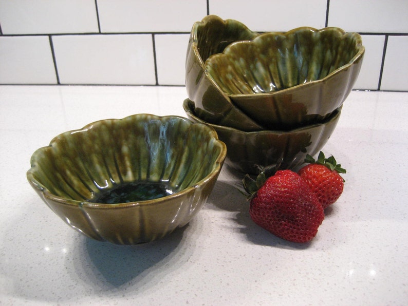 May include: Set of four green ceramic bowls with a scalloped edge. The bowls are stacked on top of each other. There are two red strawberries in the foreground.