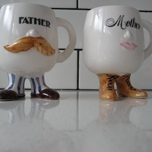 May include: Two white ceramic mugs with painted faces and legs. The mug on the left has a brown mustache and the text "FATHER" on the front. The mug on the right has pink lips and the text "Mother" on the front.