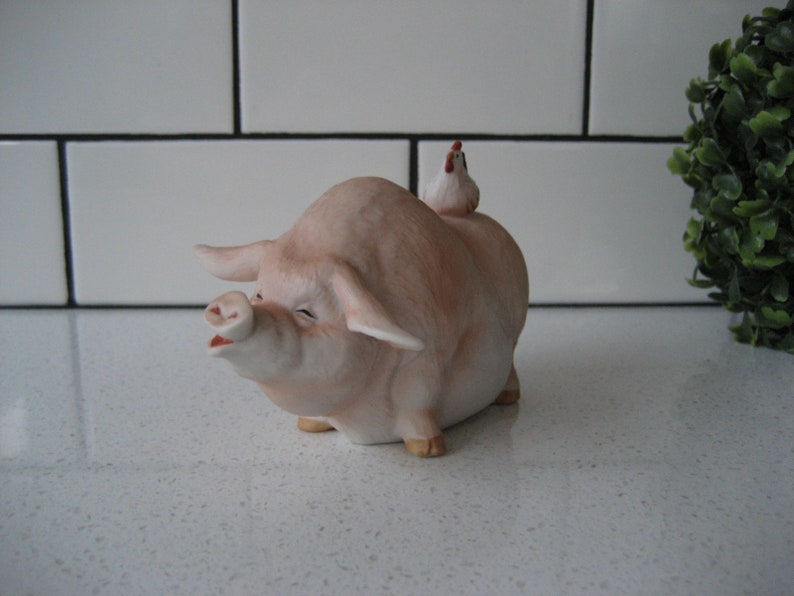 May include: A ceramic figurine of a pink pig with a small white chicken perched on its back. The pig has a smiling expression with its mouth open. The figurine is a decorative item.