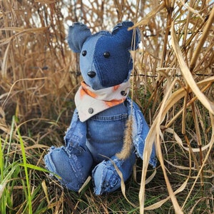 May include: A denim teddy bear with a blue and white patchwork design, wearing an orange and white bandana. The bear is sitting in a field of tall grass.