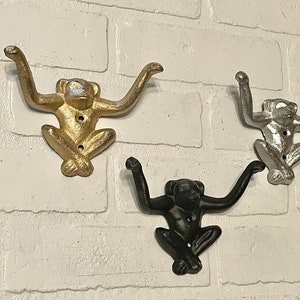 Può includere: Tre ganci a forma di scimmia in oro, argento e nero. I ganci sono fissati a un muro di mattoni bianchi.