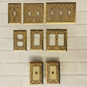 以下が含まれることがあります： 装飾的な金色のライトスイッチとコンセントカバーのコレクション。カバーには、複雑な花柄と幾何学模様が施されています。シングル、ダブル、トリプルスイッチプレート、コンセントカバーなど、さまざまなサイズと構成が表示されています。