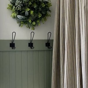 May include: A decorative green wreath with various leaves and succulents hangs above three black metal hooks on a sage green wall. A striped curtain is on the right, and a window with a stained-glass design is on the left.
