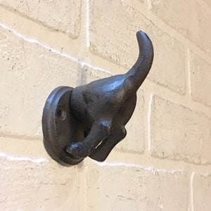May include: A black cast iron dog hook with a curled tail. The hook is mounted on a white brick wall.