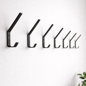 May include: A row of six matte black metal wall hooks mounted in a horizontal line. Each hook features a flat, angled design with two visible screw holes on the vertical mounting plate and a curved upward-facing prong for hanging items.