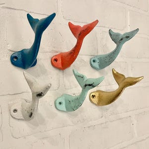 Może przedstawiać: Sześć haczyków w kształcie ogona wieloryba wykonanych z żeliwa w różnych kolorach, w tym turkusowym, białym, złotym, koralowym i niebieskim. Haczyki są zamontowane na białej ceglanej ścianie.