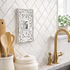 Puede incluir: Interruptor de luz blanco y ornamentado con un acabado envejecido, montado en un salpicadero de azulejos blancos. El interruptor está rodeado por un diseño floral. También se ven un grifo dorado y un azucarero.