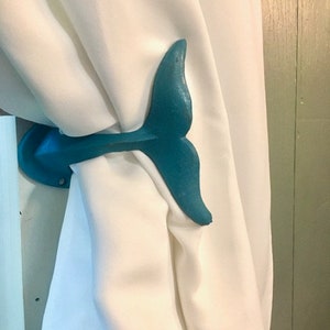 May include: Teal blue whale tail curtain tieback. The tieback is made of metal and is shaped like a whale tail. It is attached to a white curtain.