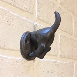 May include: A black cast iron dog hook mounted on a white brick wall. The dog is in a running position with its tail curled up.