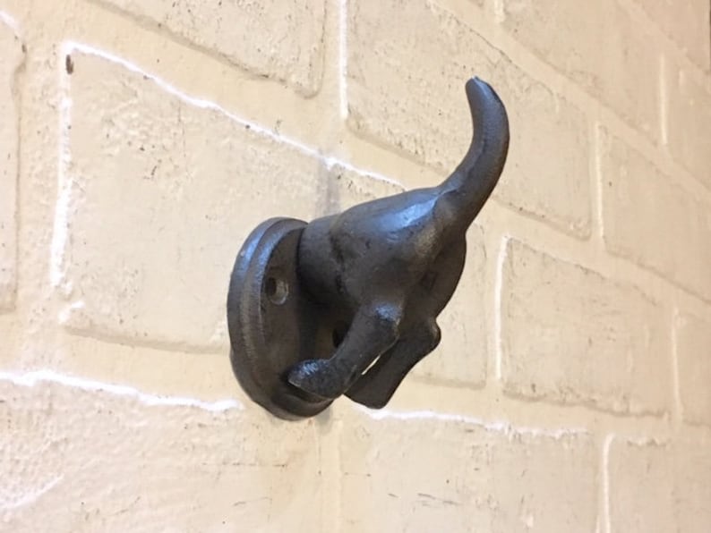 May include: A black cast iron dog hook mounted on a white brick wall. The dog is in a running position with its tail curled up.
