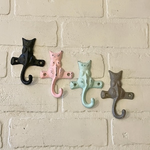 May include: Four cast iron cat-shaped wall hooks in black, pink, mint green, and brown. Each hook has a curved tail and a mounting plate with two holes for screws. The hooks are mounted on a white brick wall.