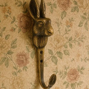 May include: A bronze-coloured metal wall hook shaped like a rabbit's head. The hook has a long, slender stem with three holes and a curved end. The background is a floral wallpaper.