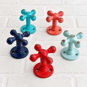 May include: Five metal wall hooks shaped like vintage faucet handles, each featuring a four-pronged cross design. The hooks are finished in distinct colors including bright red, dark blue, turquoise, coral, and light blue, all with distressed edges.