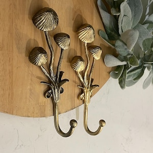 Puede incluir: Dos ganchos decorativos de pared con diseño de seta. Los ganchos son de metal y tienen un acabado dorado y bronce. Los sombreros de las setas tienen un diseño texturizado y acanalado. Los ganchos están montados en una placa de madera.