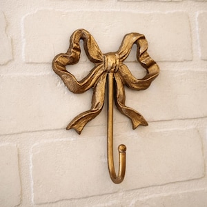 May include: A gold-coloured wall hook featuring a decorative bow design at the top. The bow has a textured, metallic finish with flowing ribbon loops and tails. A single, smooth, curved hook extends downwards from the centre of the bow structure.