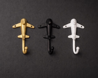 Può includere: Tre ganci da parete a forma di aeroplano in oro, nero e bianco. Ogni gancio ha un fondo curvo per appendere oggetti. I ganci sono in metallo e sono posizionati su uno sfondo scuro.