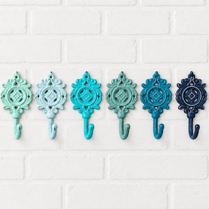May include: A set of six wall hooks arranged in a horizontal row, each featuring an ornate, symmetrical metal design with a central circular motif. The hooks display a gradient of blue and green shades, ranging from light mint to deep navy blue.
