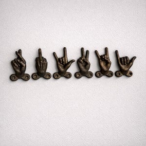 Puede incluir: Seis ganchos de pared con forma de mano de color bronce, cada uno representando un gesto diferente. Los ganchos están montados sobre pequeñas bases decorativas con agujeros para tornillos. Los gestos incluyen dedos cruzados, dedo medio, rock on, signo de la paz y shaka.