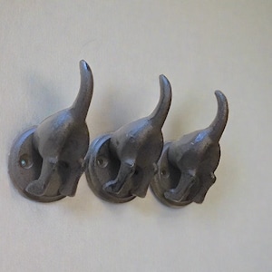 May include: Three black metal dog-shaped hooks mounted on a white wall. The hooks are in a row and are facing the same direction. Each hook has a rounded base and a dog's head with a tail that curves up.