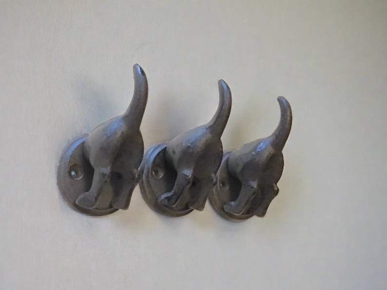 May include: Three black metal dog-shaped hooks mounted on a white wall. The hooks are in a row and are facing the same direction. Each hook has a rounded base and a dog's head with a tail that curves up.