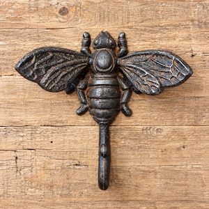 May include: A dark, weathered cast iron wall hook shaped like a bee. The insect features detailed, textured wings and a segmented body, with the lower portion of the body extending downward into a functional, curved hook for hanging items.