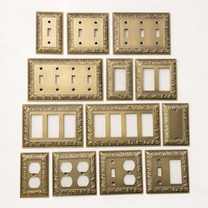 Puede incluir: Una colección de placas de interruptores de luz de color latón antiguo en varias configuraciones. Las placas presentan patrones ornamentados y arremolinados alrededor de los bordes. Los estilos incluyen placas de interruptores simples, dobles, triples y cuádruples, junto con cubiertas de enchufes.