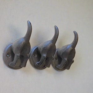 May include: Three black metal dog-shaped hooks mounted on a white wall. The hooks are in a row and are facing the same direction. Each hook has a rounded base and a dog's head with a long tail that curves up and over the head.