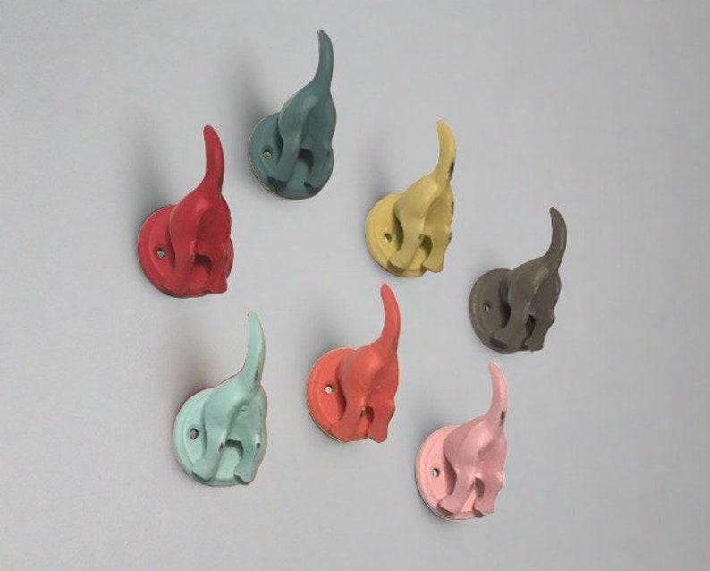May include: Seven colorful cat-shaped hooks, each with a different color: teal, red, yellow, gray, light blue, coral, and pink. The hooks are mounted on a white wall.