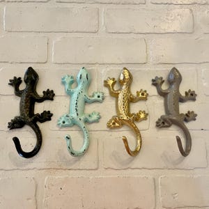 May include: Four lizard-shaped wall hooks in black, light blue, gold, and grey. Each hook is made of metal and has a curved tail for hanging items. The hooks are mounted on a white brick wall.