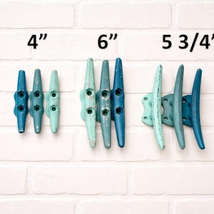 May include: Three sets of blue and teal boat cleats, 4", 6", and 5 3/4" long, mounted on a white brick wall. The cleats are in varying shades of blue and teal, with a distressed finish. The image shows the cleats' dimensions.