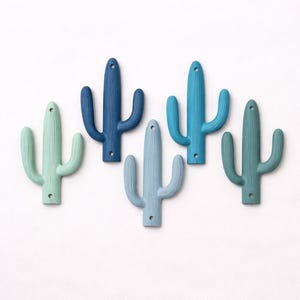 Pode incluir: Cinco ganchos de parede em forma de cacto em tons de azul e verde. Cada gancho tem um furo na parte superior para pendurar e um furo na parte inferior. Os ganchos estão dispostos sobre um fundo branco.