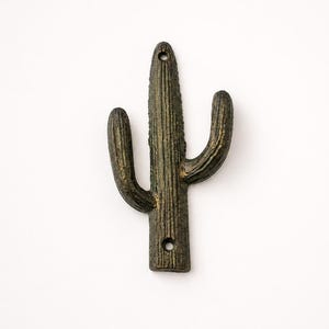 May include: A dark bronze-colored metal wall hook shaped like a saguaro cactus. The surface features a textured, ribbed finish with two circular mounting holes, one located at the top and one at the bottom of the central vertical column.