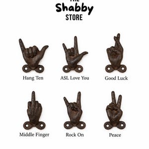 Op de afbeelding: Een verzameling van zes donkerbruine gietijzeren wandhaken in de vorm van handen met verschillende gebaren. De tekst THE Shabby STORE staat bovenaan, met de labels Hang Ten, ASL Love You, Good Luck, Middle Finger, Rock On en Peace onder elke haak.