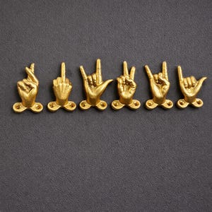 Può includere: Un set di sei ganci da parete in metallo color oro, ognuno a forma di mano che mostra un gesto diverso. Le mani sono disposte in una fila orizzontale, con dita e nocche dettagliate, e ogni base include due fori per le viti per il fissaggio.