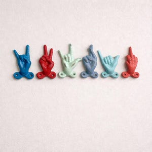 Puede incluir: Un conjunto de seis colgadores de pared con forma de gestos de manos, en colores vibrantes. Cada colgador es de un color diferente: azul, rojo, verde claro, azul grisáceo, azul claro y rojo. Montados sobre una superficie blanca.