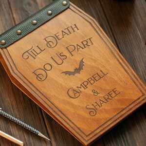 Puede incluir: Una caja de madera con forma de ata&uacute;d con una correa de cuero negro y tachuelas met&aacute;licas. La caja est&aacute; grabada con el texto "Till Death Do Us Part" y los nombres "Campbell & Sharee".