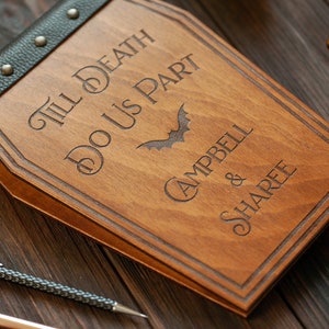 Puede incluir: Un libro de visitas de madera con forma de ata&uacute;d con una cubierta de cuero negro y tachuelas met&aacute;licas. El libro est&aacute; grabado con el texto "Till Death Do Us Part" y un gr&aacute;fico de murci&eacute;lago. Los nombres "Campbell & Sharee" tambi&eacute;n est&aacute;n grabados en el libro.